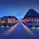 A wooden walkway with deck lighting leads to two thatch-roofed, overwater bungalows at dusk. Warm interior lights contrast with the deep blue ocean and sky, creating a tranquil, inviting scene as the structures stand on stilts over calm water.